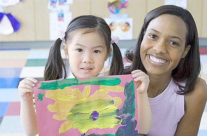 A girl sitting beside her teacher in the classroom holding painting. Net photo.